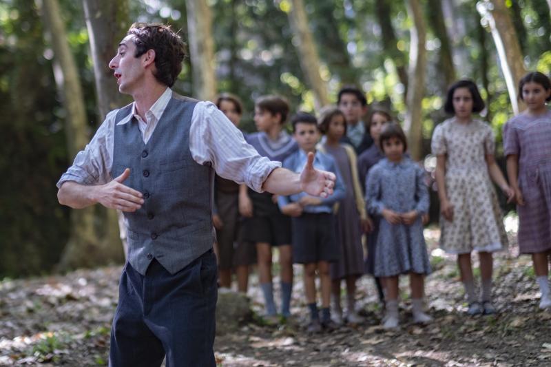 Filmszene aus "Der Lehrer, der uns das Meer versprach". Ein junger Mann steht gestikulierend vor einer Schar Kinder in einem Wald. c) Filmax 