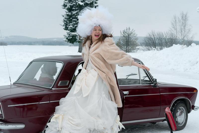 Filmszene aus "Neuigkeiten aus Lappland". Eine Frau im Hochzeitskleid steigt aus einem roten Auto mitten im Schnee. 