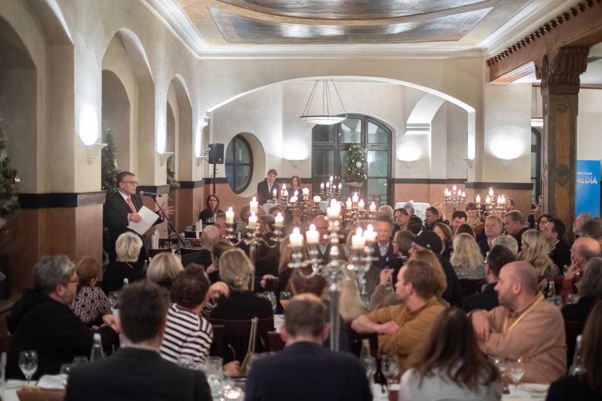 Ein Saal voller Menschen an Tischen, hell erleuchtet, hören einem Mann auf einem Podes zu.