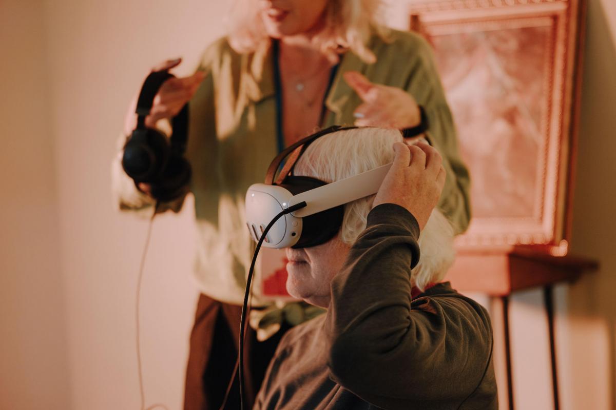 Eine Person setzt eine VR Brille auf, eine andere hilft ihr dabei.