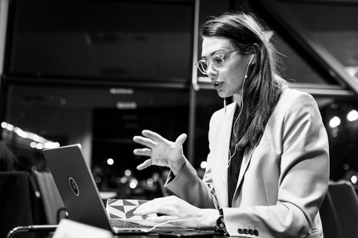 eine Frau spricht in den Laptop und hat Kopfhörer im Ohr, sie gestikuliert dabei.