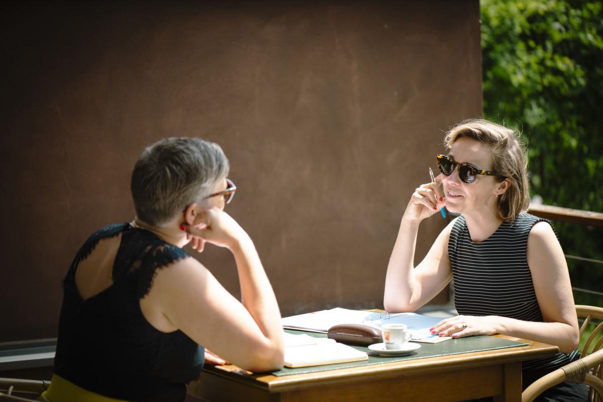 zwei Frauen sitzen sich bei Sonnenschein gegenüber und lächeln sich an. 